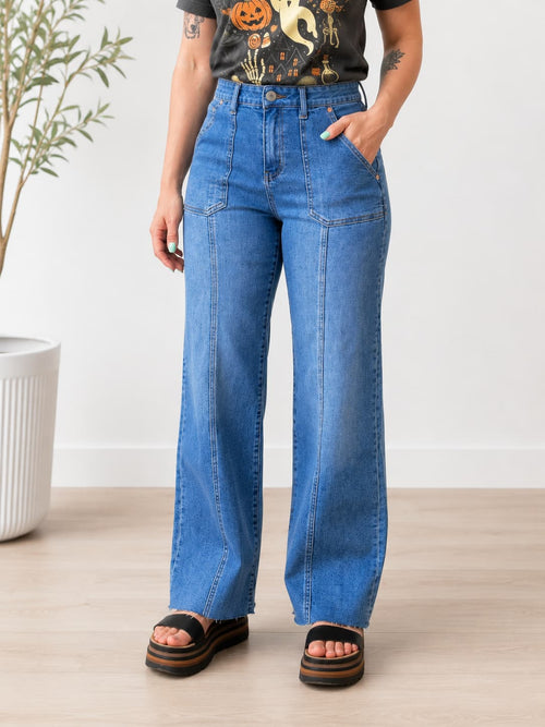 A person wearing high-waisted, wide-leg blue denim jeans with a black graphic t-shirt and black platform sandals, standing in a brightly lit room with a light wood floor and a white wall.