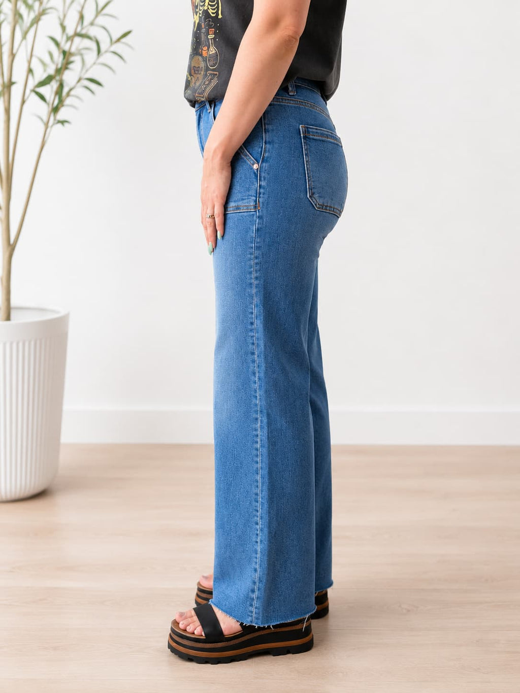 A person shown from the waist down wearing blue wide-leg flare jeans, a black graphic t-shirt, and black platform sandals in a bright room with a light wood floor and a potted plant.