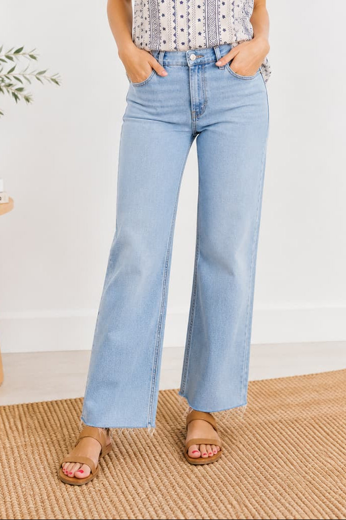 Woman wearing a patterned blouse and light blue jeans standing in a room with a white wall and wooden side table.