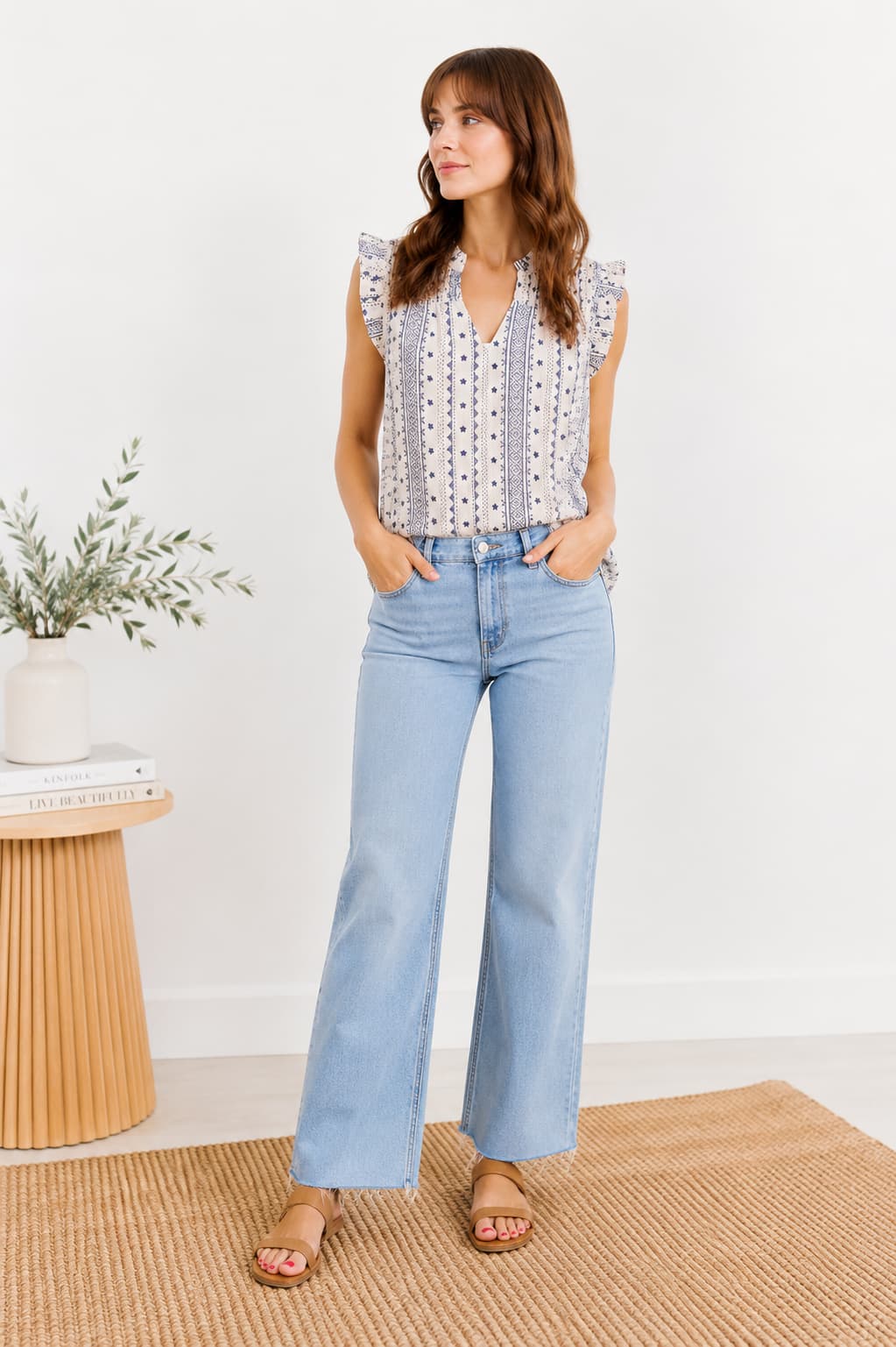 Woman wearing a patterned blouse and light blue jeans standing in a room with a white wall and wooden side table.