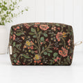 A rectangular cosmetic pouch with a dark brown background and a detailed floral print in pink, green, and yellow. It features a silver zipper on the left with a small white tag, and a beige loop handle on the right, set against a white paneled background.