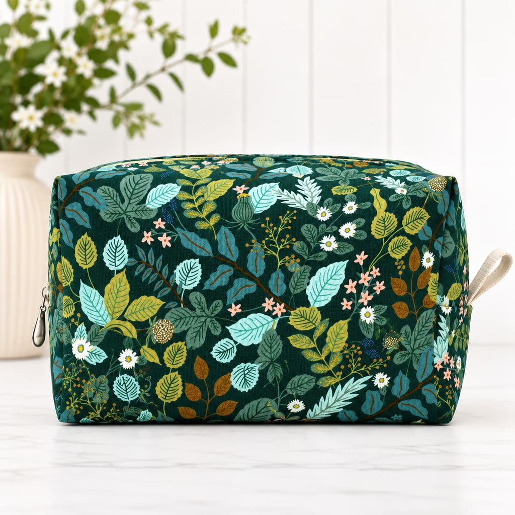 A rectangular zippered pouch with a dark green background and an all-over botanical print featuring teal, yellow, and white leaves and small flowers, shown on a light surface with a blurred plant in the background.