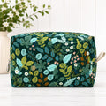 A rectangular zippered pouch with a dark green background and an all-over botanical print featuring teal, yellow, and white leaves and small flowers, shown on a light surface with a blurred plant in the background.