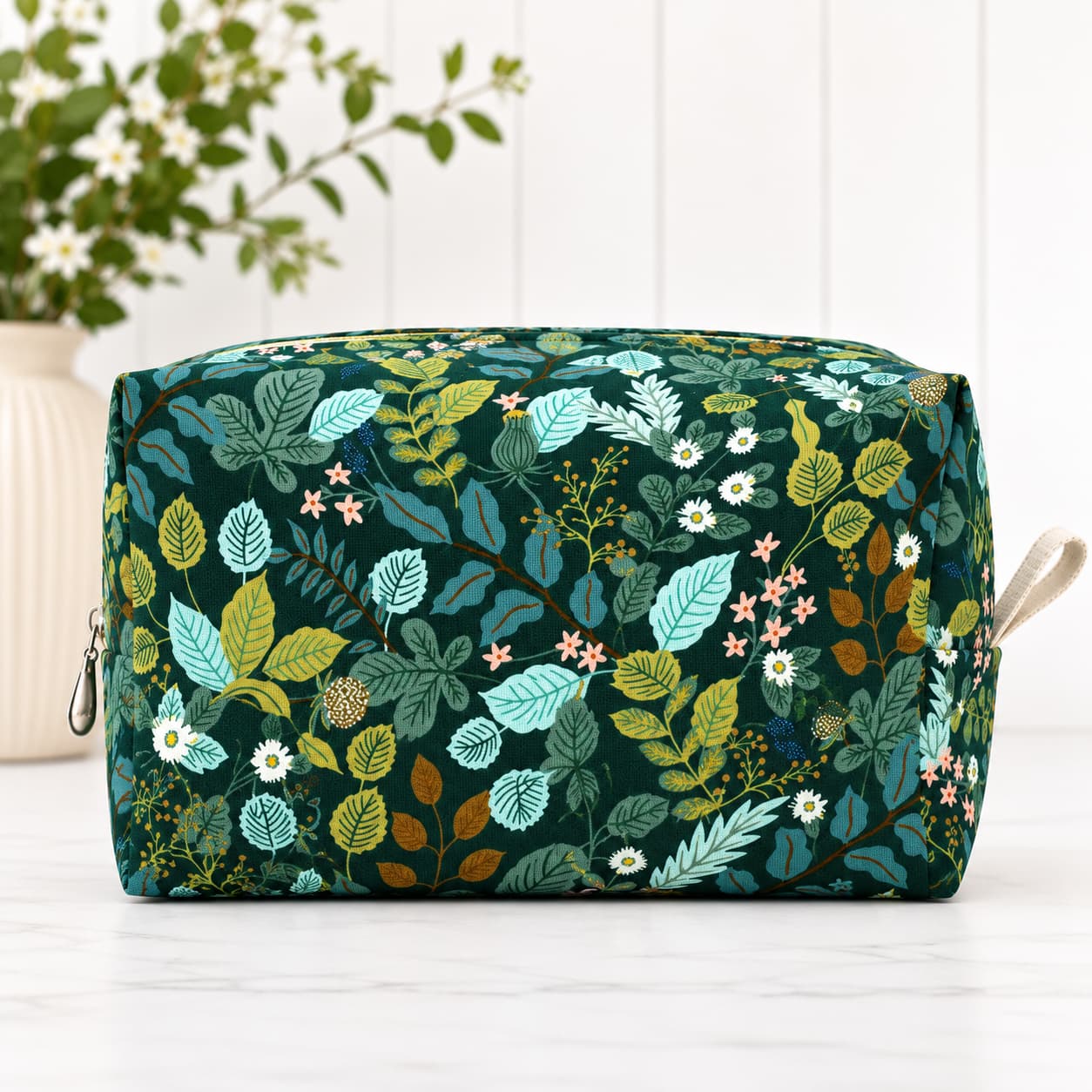 A rectangular zippered pouch with a dark green background and an all-over botanical print featuring teal, yellow, and white leaves and small flowers, shown on a light surface with a blurred plant in the background.