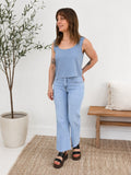 Woman wearing a blue sleeveless top and jeans standing in a room with a plant and wooden bench.