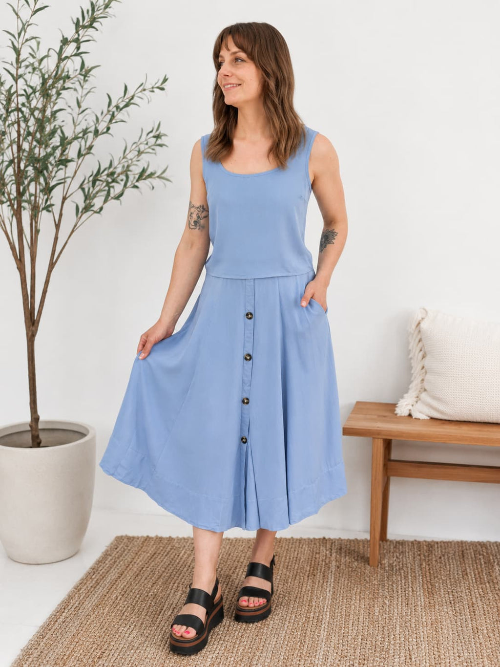 Woman wearing a two piece blue dress standing in a room with a plant and wooden bench.