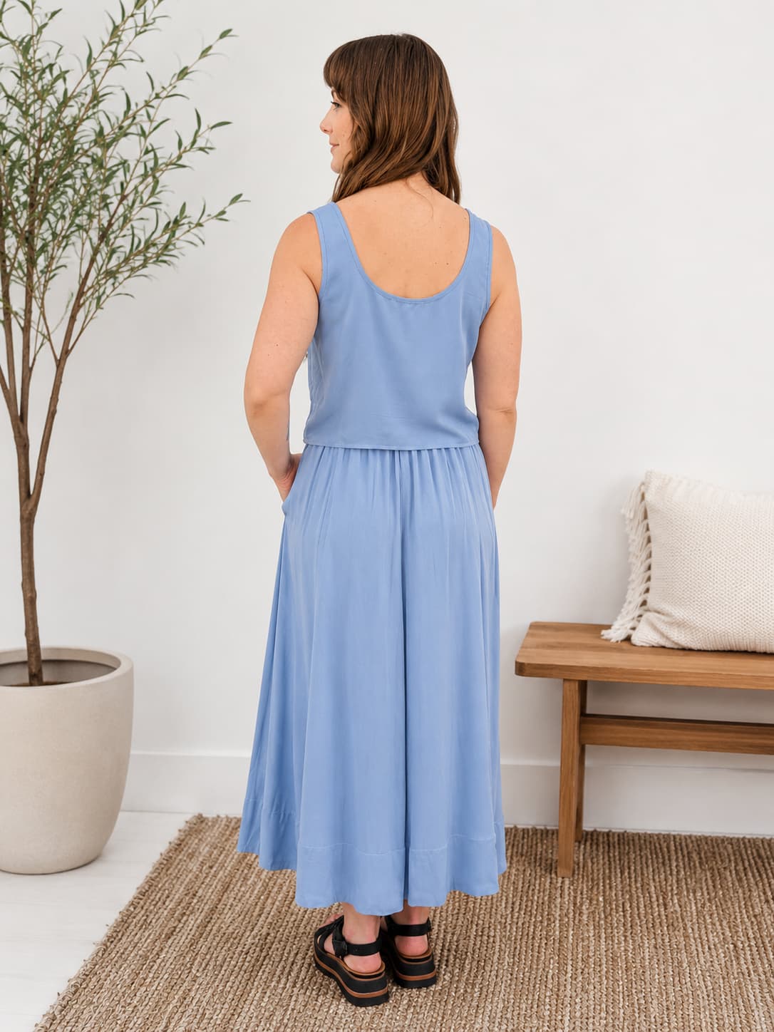 Woman wearing a two piece light blue dress standing in a room with a plant and wooden bench.