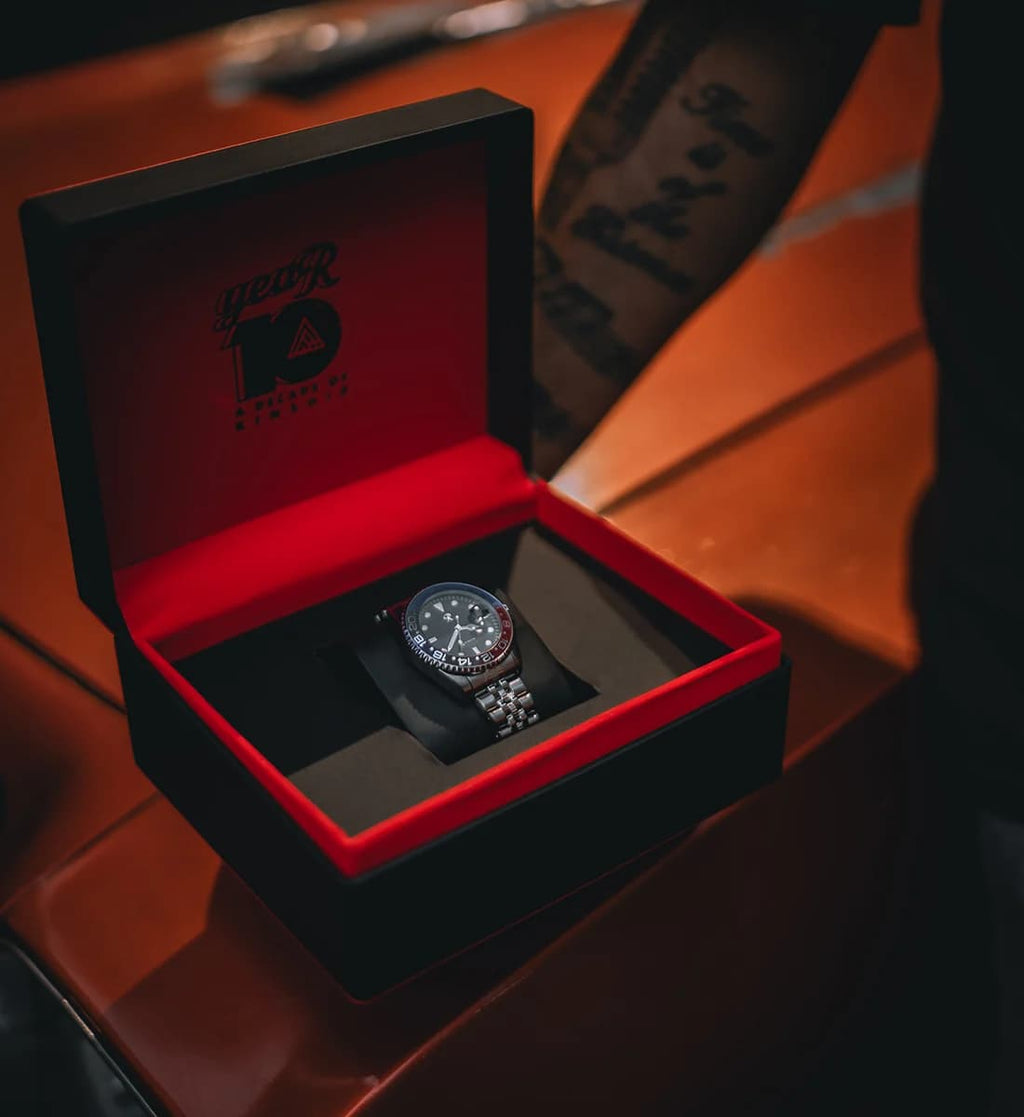 A silver-toned wristwatch with a dark dial and metal link band resting inside an open black presentation box with a red interior. The box lid is shown with a red interior and black printed text/logo, and the scene is photographed on a warm brown surface with a blurred tattooed arm in the background.