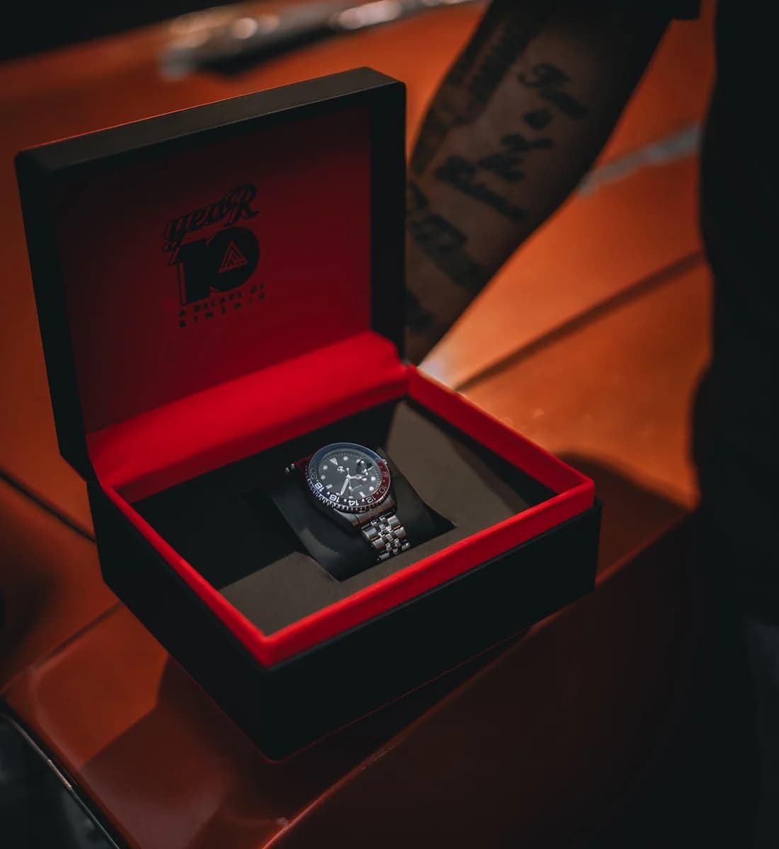 A silver-toned wristwatch with a dark dial and metal link band resting inside an open black presentation box with a red interior. The box lid is shown with a red interior and black printed text/logo, and the scene is photographed on a warm brown surface with a blurred tattooed arm in the background.