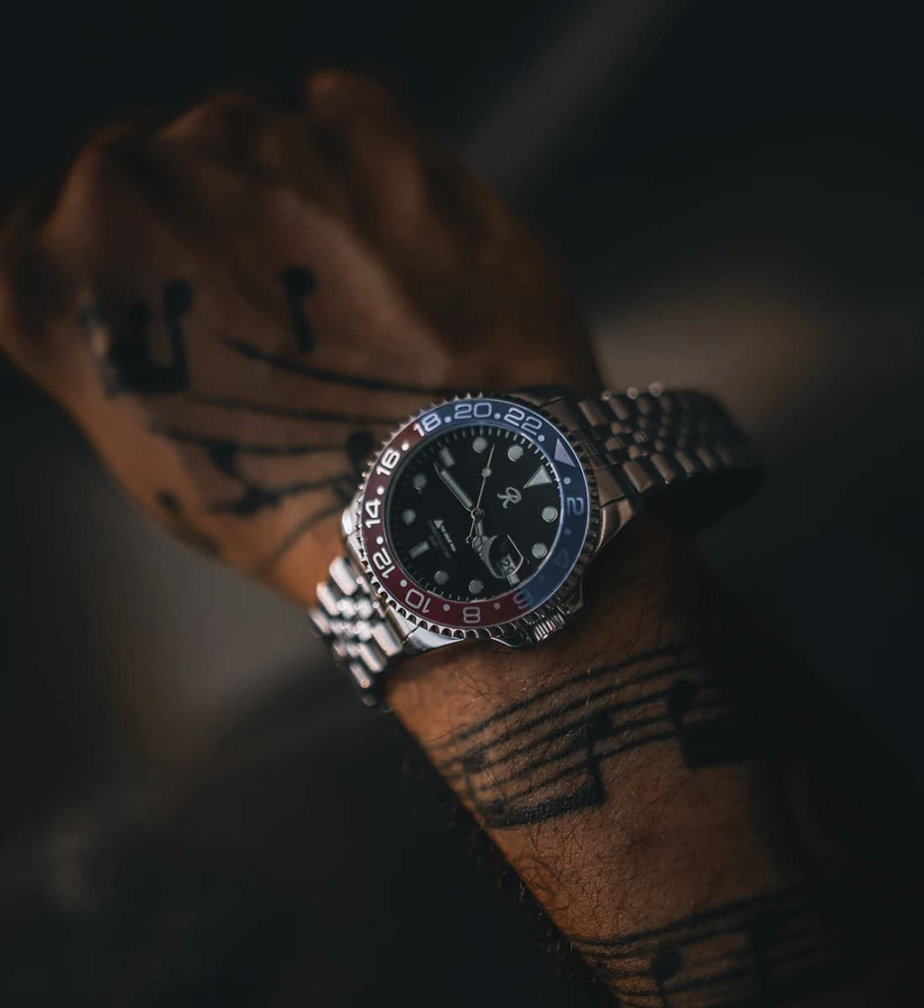 A wristwatch with a silver metal link bracelet, a black dial, and a red and blue bezel, shown worn on a tattooed wrist against a dark background.