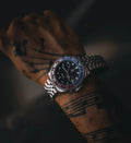 A wristwatch with a silver metal link bracelet, a black dial, and a red and blue bezel, shown worn on a tattooed wrist against a dark background.