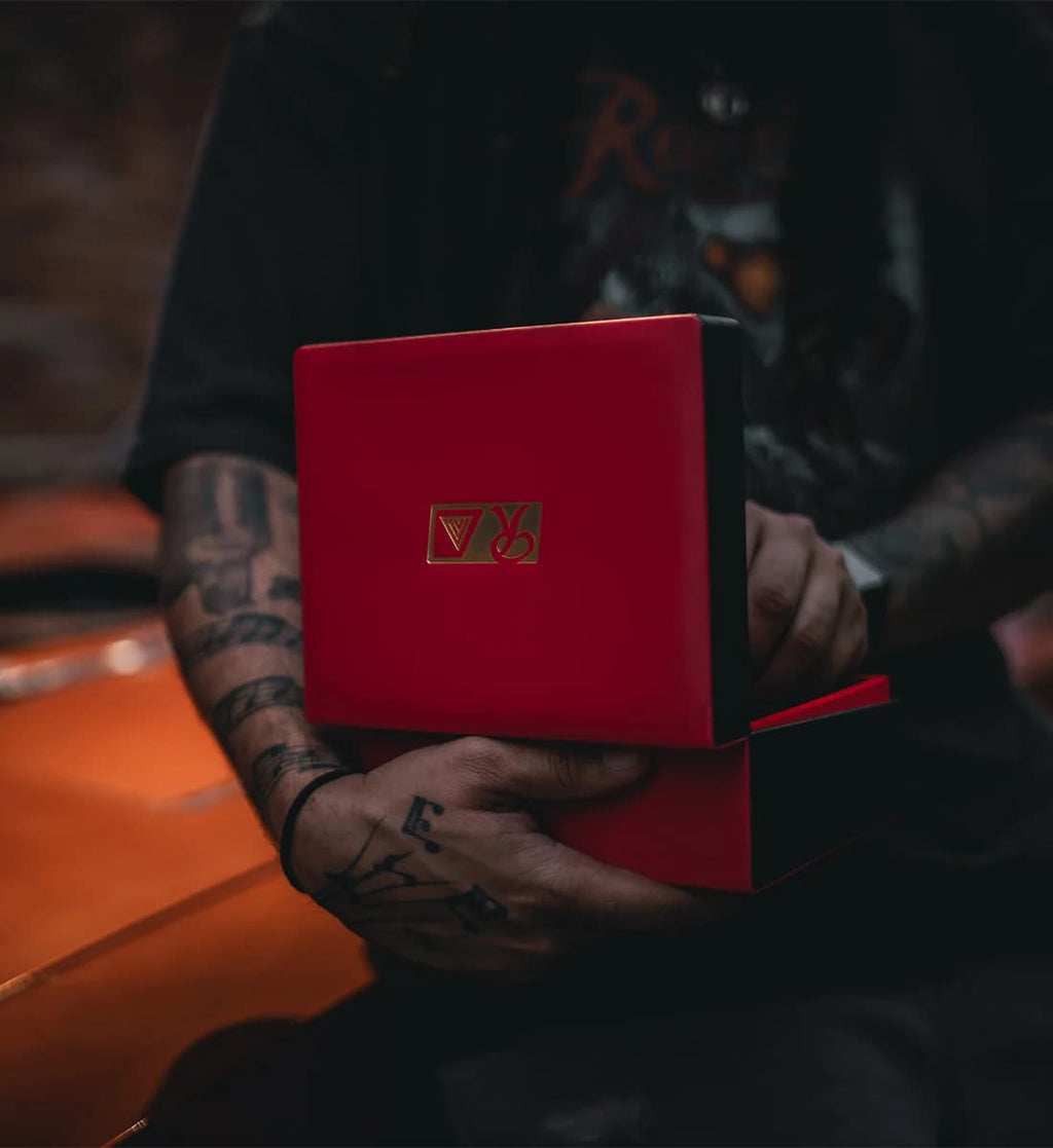 A person with tattooed arms holds an open red presentation box with a gold and red logo on the lid. The person is wearing a black graphic t-shirt, and the background is dark with a blurred orange surface in the foreground.
