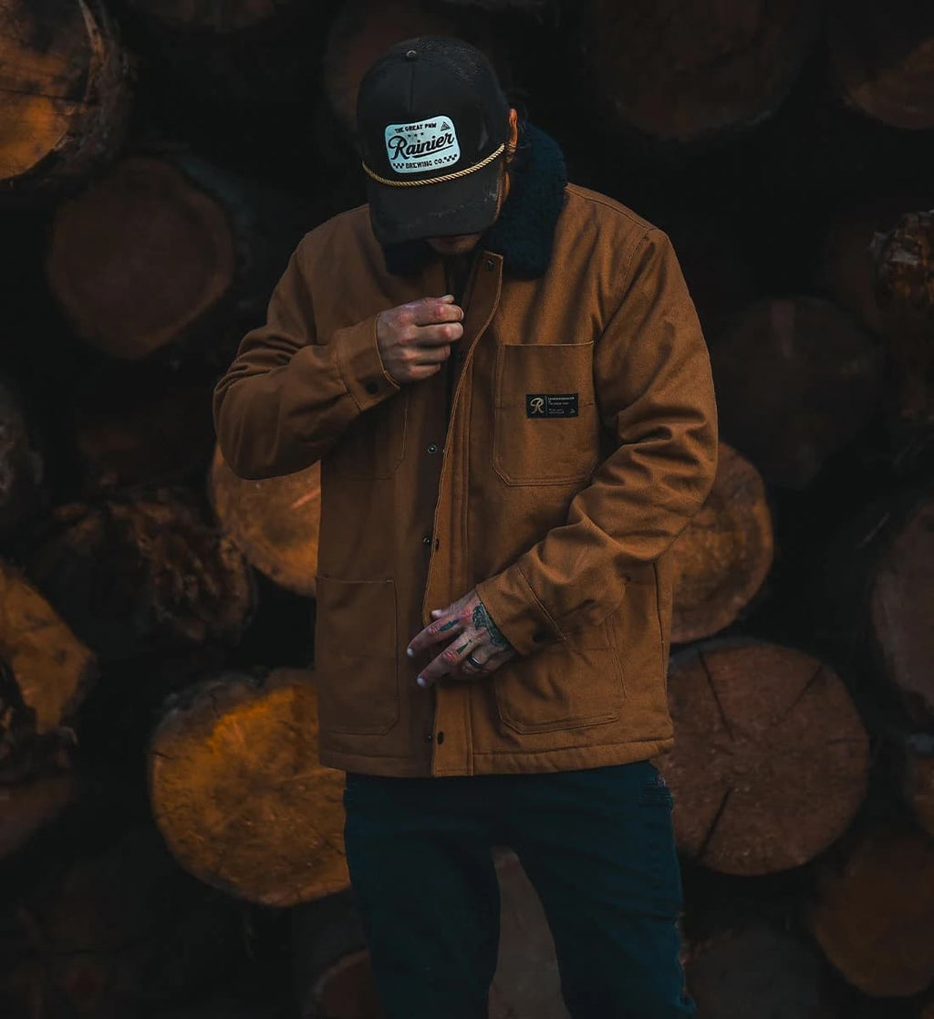 A man wearing a brown Carhartt jacket with a black fleece collar, a black trucker hat with a 'Rustico' patch, and dark pants, standing in front of a stack of cut logs.