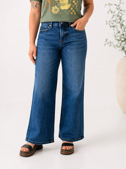 A person wearing medium-wash blue wide-leg denim jeans, paired with a green graphic t-shirt and brown slide sandals, standing against a clean white studio background.