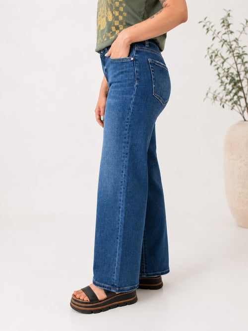 A person shown from the waist down wearing blue wide-leg jeans, a green patterned top, and black platform sandals, standing against a clean white studio background with a plant in a vase at the side.