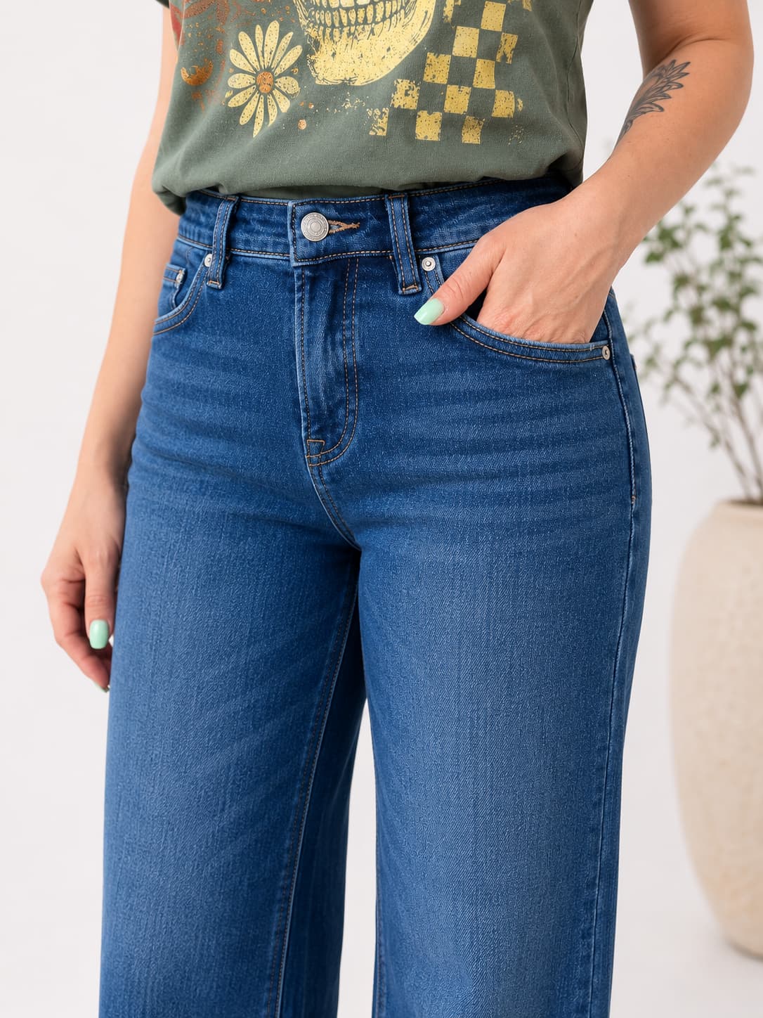 A person wearing medium-wash blue denim jeans with a hand in the pocket, paired with an olive green graphic t-shirt. The photo is cropped from the chest down against a clean white background with a blurred potted plant on the right.