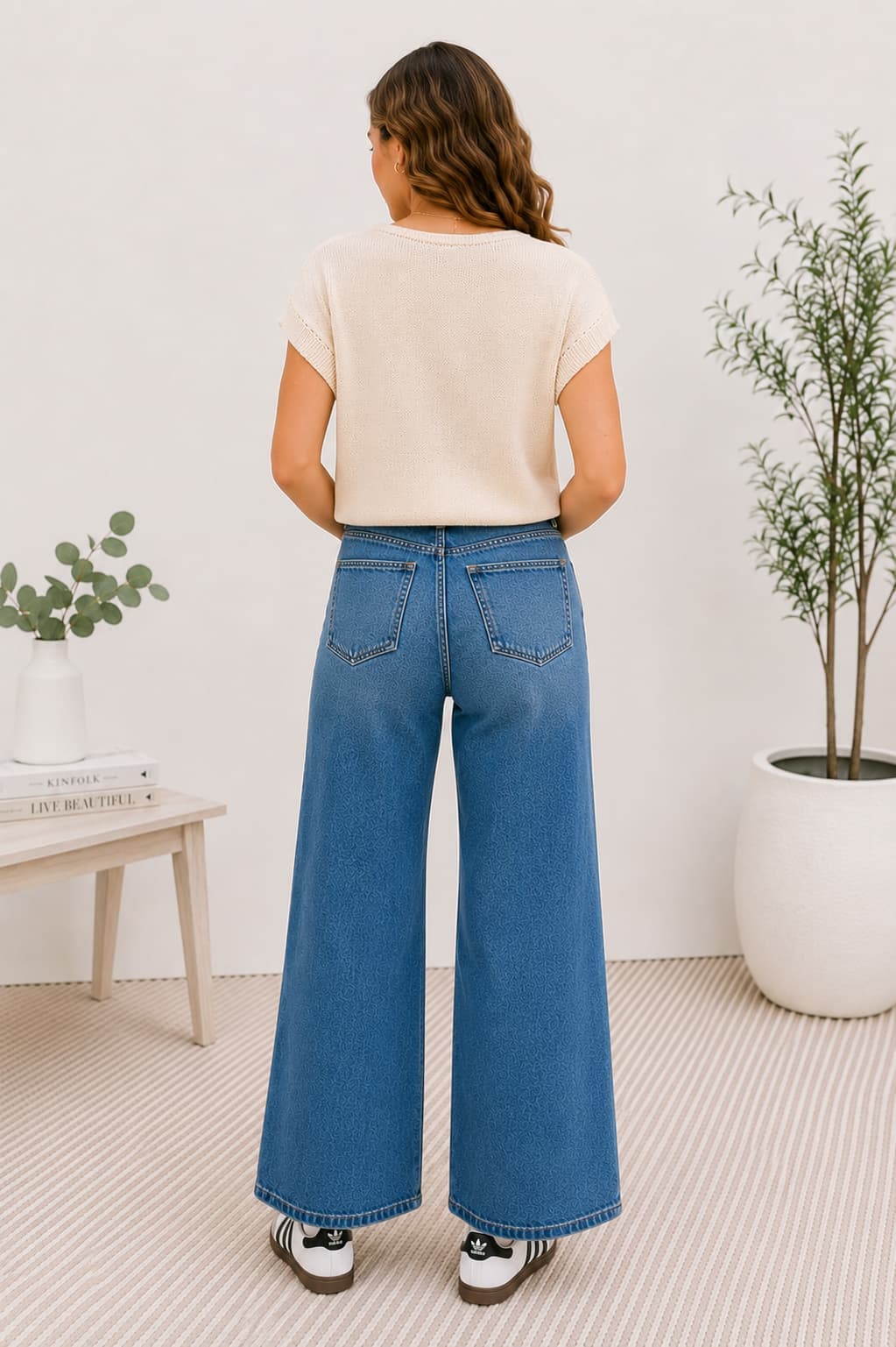 Person wearing blue wide-leg jeans and a beige top in a room with plants and a table.