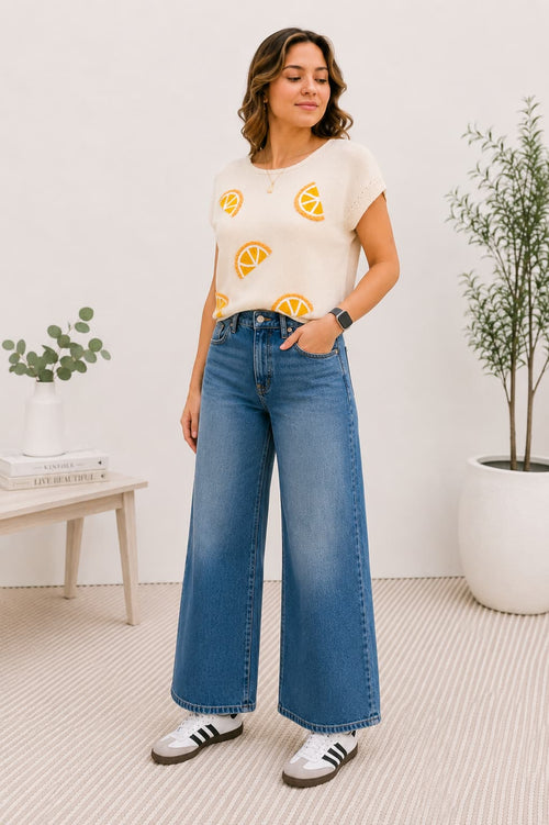 Woman wearing a knit top with orange designs and blue jeans indoors.