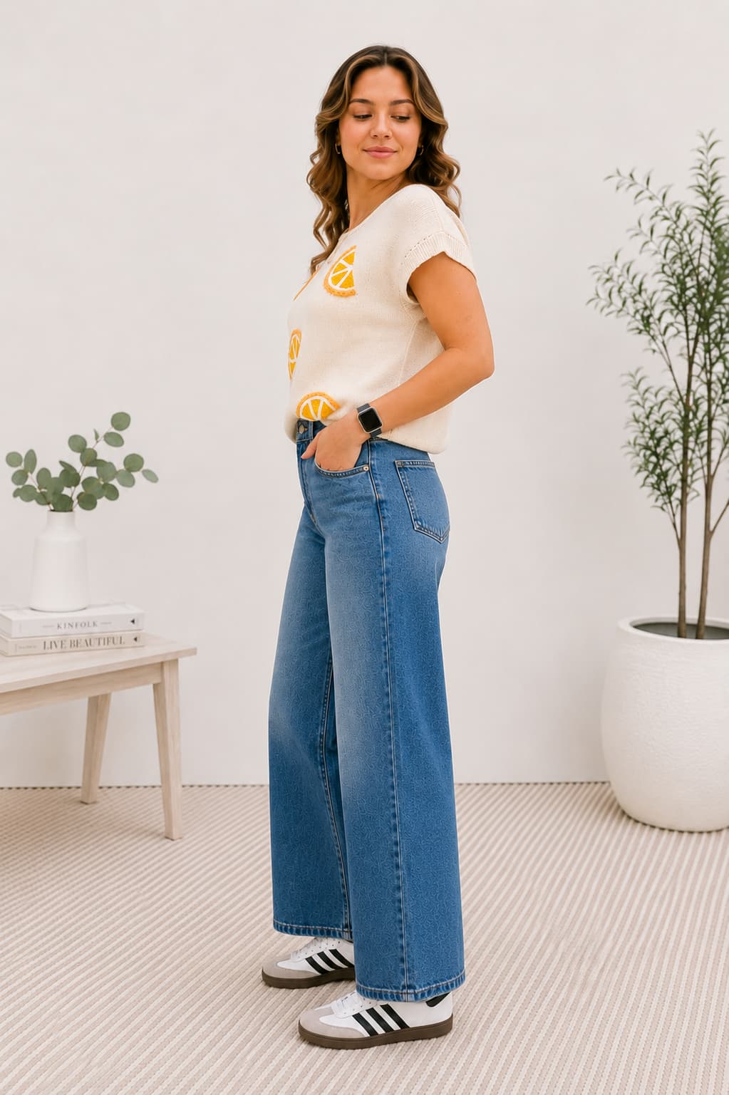 Woman wearing a knit top with oranges and blue jeans in a room with plants and a table.