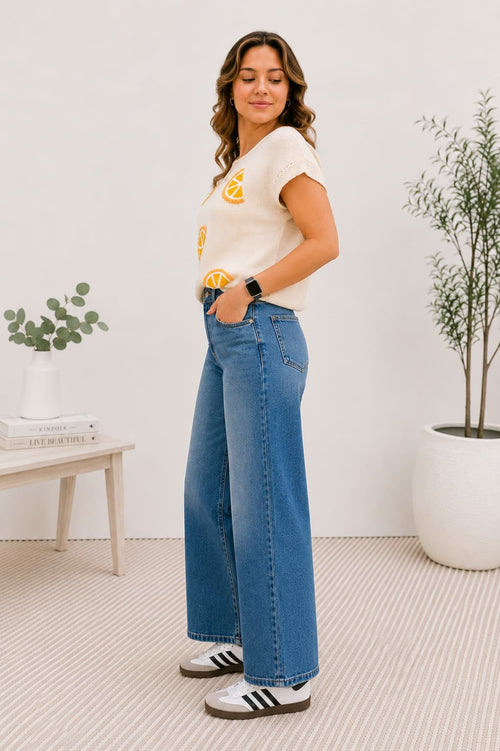 Woman wearing a knit top with oranges and blue jeans in a room with plants and a table.
