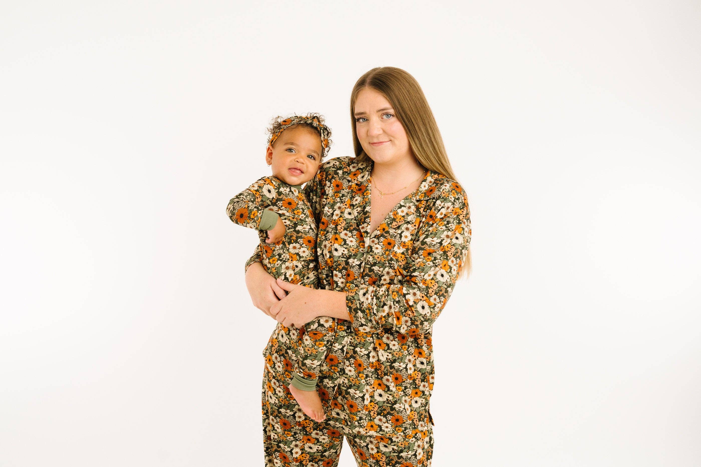 A woman holding a baby, both wearing matching floral print long-sleeve pajamas and a matching headband, against a plain white background.