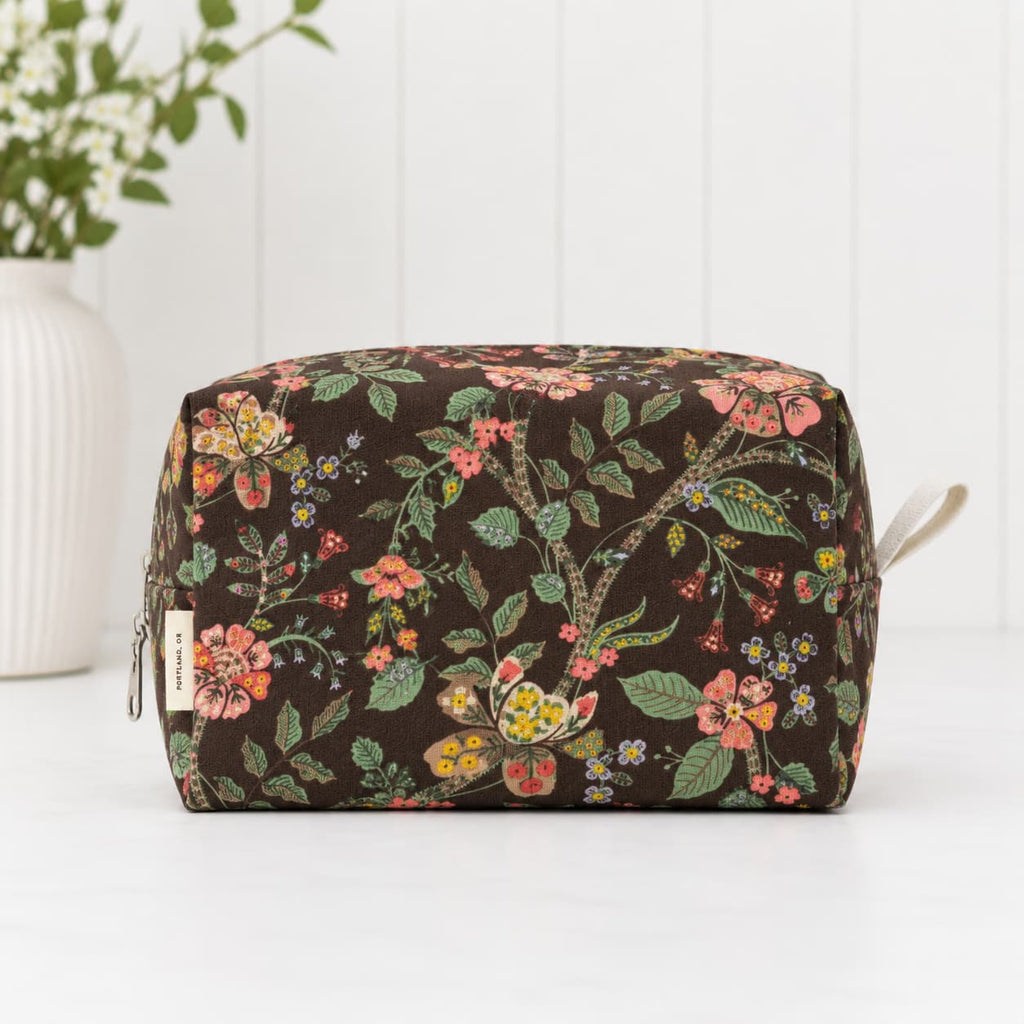 A rectangular zippered pouch with a dark brown background and a colorful floral print, featuring a small fabric brand tag on the left side and a light-colored zipper pull on the right, set against a white paneled background.