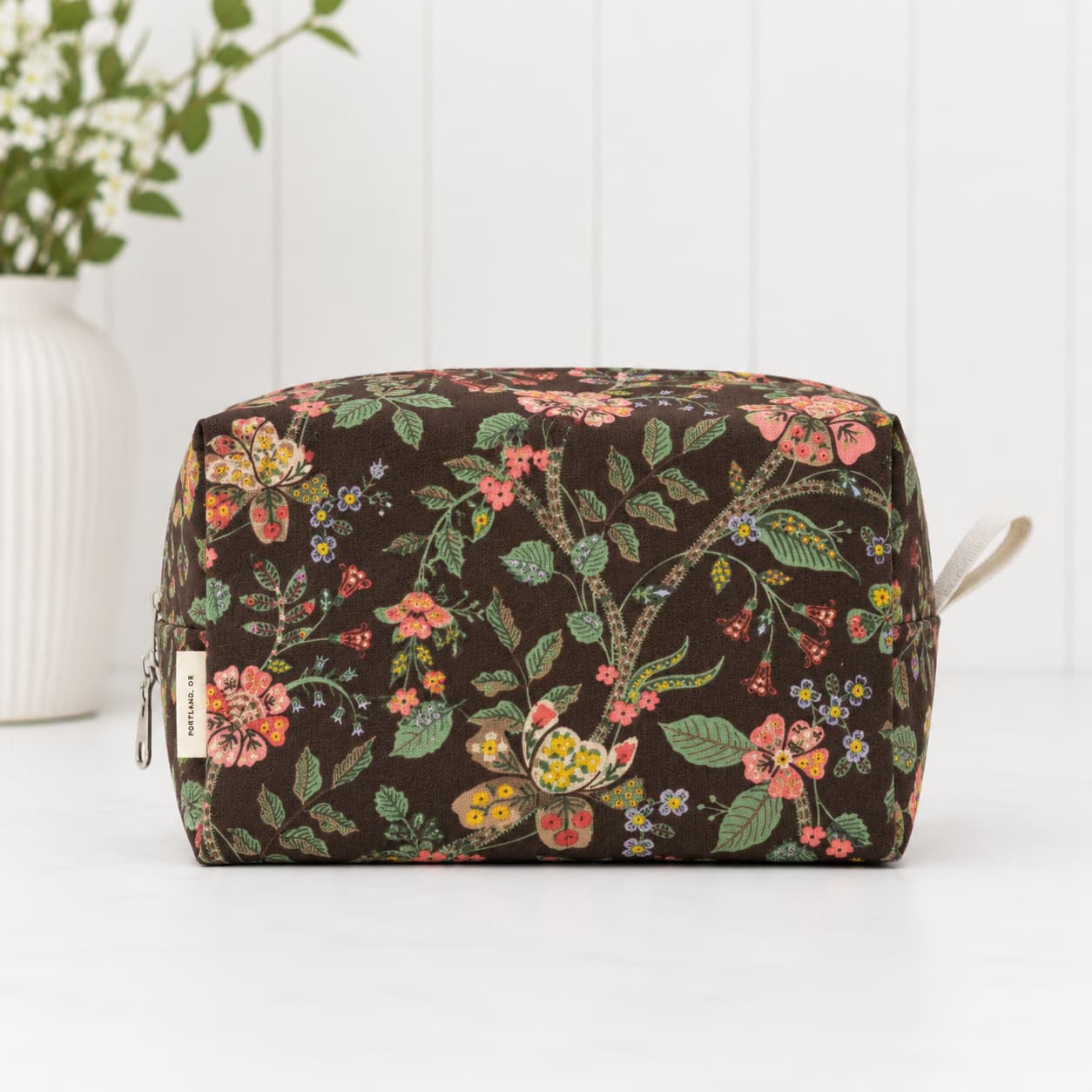 A rectangular zippered pouch with a dark brown background and a colorful floral print, featuring a small fabric brand tag on the left side and a light-colored zipper pull on the right, set against a white paneled background.