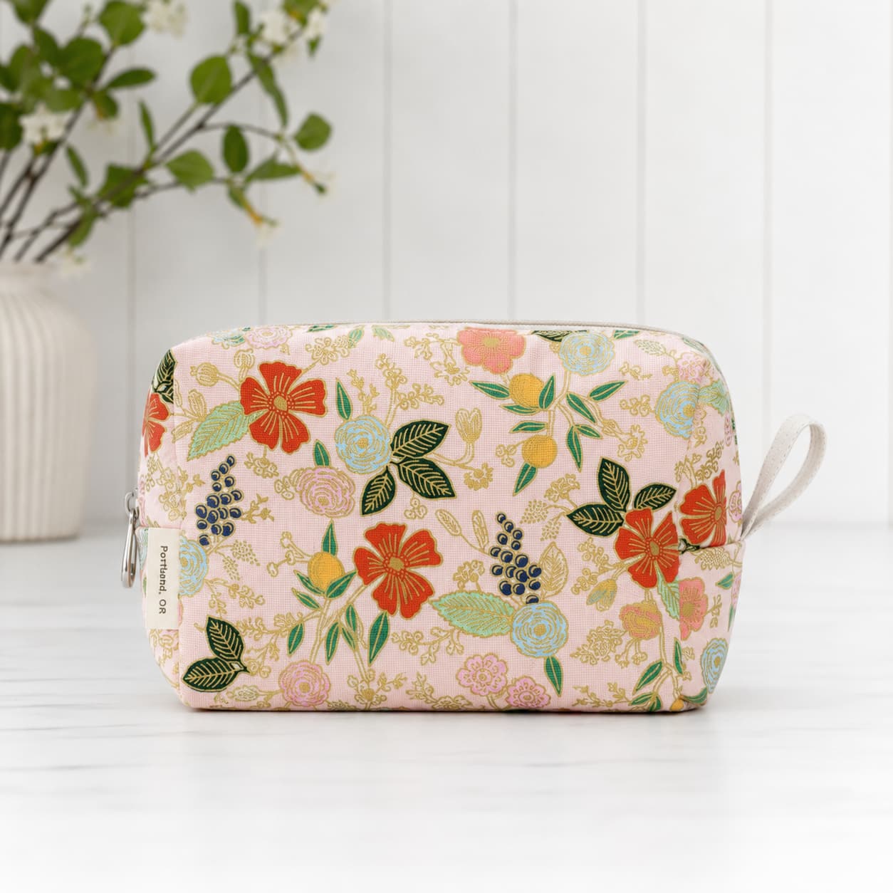 A rectangular zippered cosmetic pouch with a light pink background and a colorful floral print, featuring red, orange, yellow, and green flowers with gold leaf accents. It has a small white loop on the right side and a zipper pull on the left.