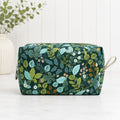 A rectangular zippered pouch with a dark green background and an all-over botanical print of leaves, flowers, and vines in shades of blue, green, yellow, and pink. It has a small beige fabric loop handle on the right side and is photographed on a light surface with a white paneled wall and a vase of greenery in the background.