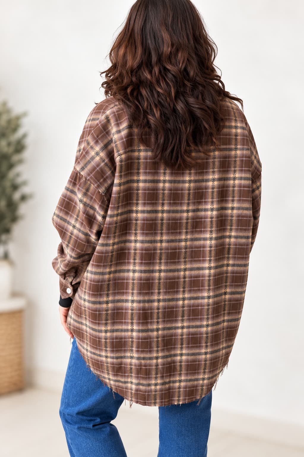 A woman viewed from behind wearing an oversized brown plaid flannel shirt with blue jeans, standing in a brightly lit indoor setting.