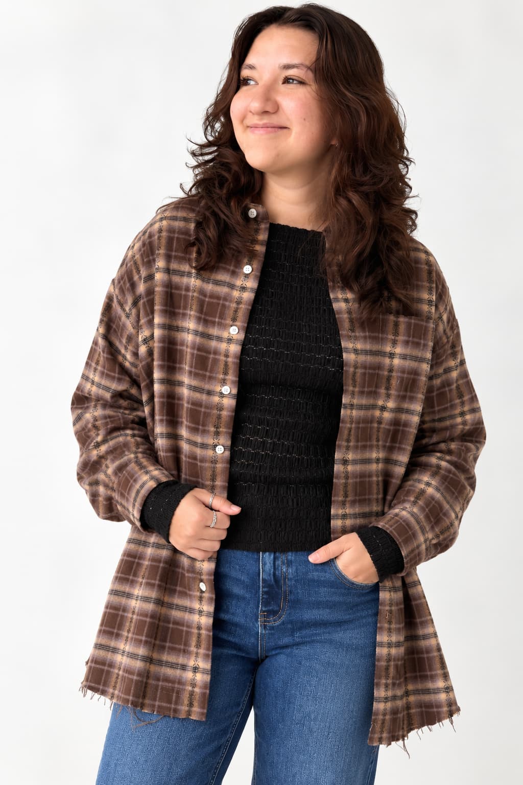 A woman wearing a brown and beige plaid button-up shirt jacket over a black knit top and blue jeans, standing against a plain white studio background.