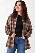 A woman wearing a brown and beige plaid button-up shirt jacket over a black knit top and blue jeans, standing against a plain white studio background.