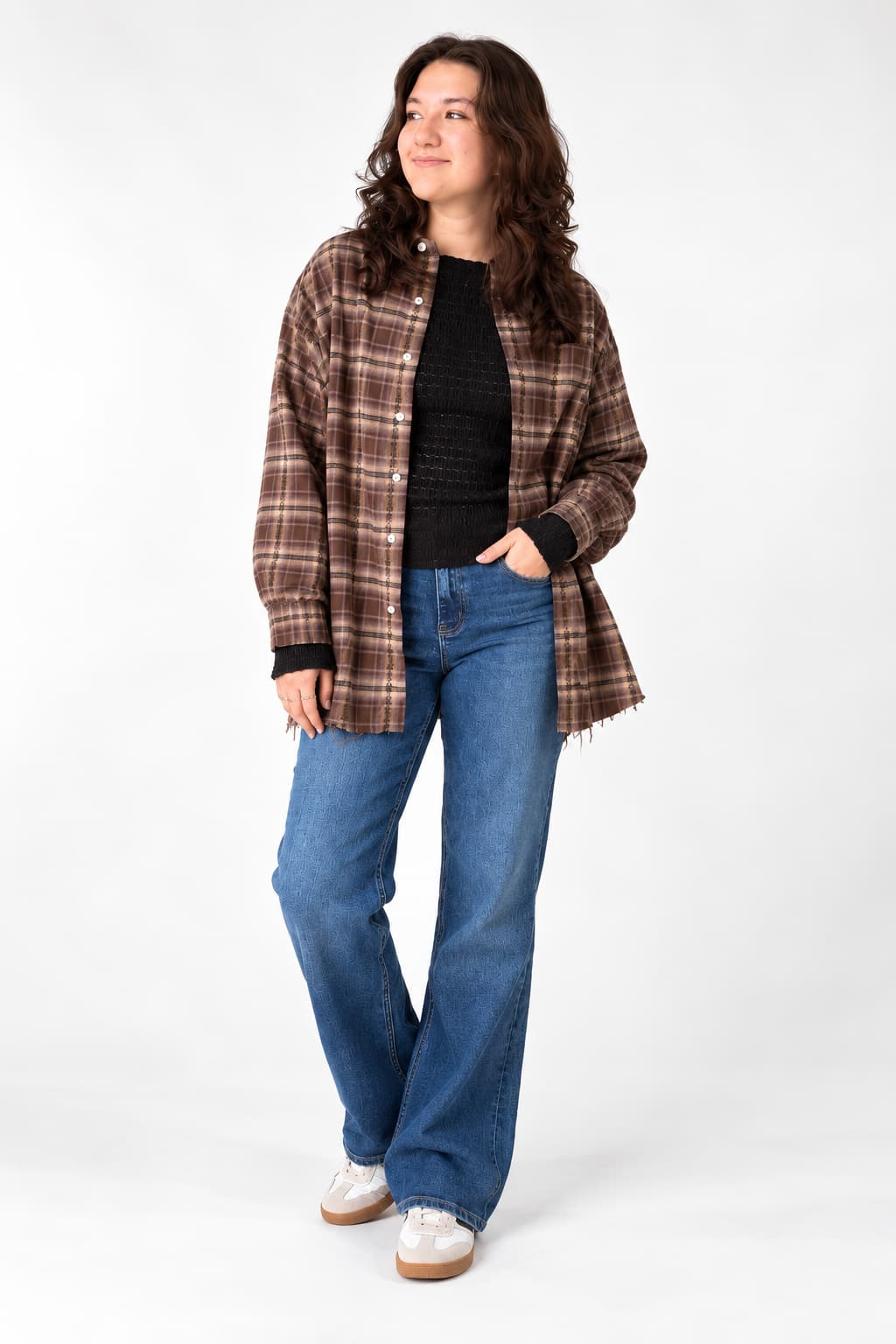 A woman modeling a brown and beige plaid button-up shirt jacket over a black ribbed top, paired with blue jeans and white sneakers, against a plain white studio background.