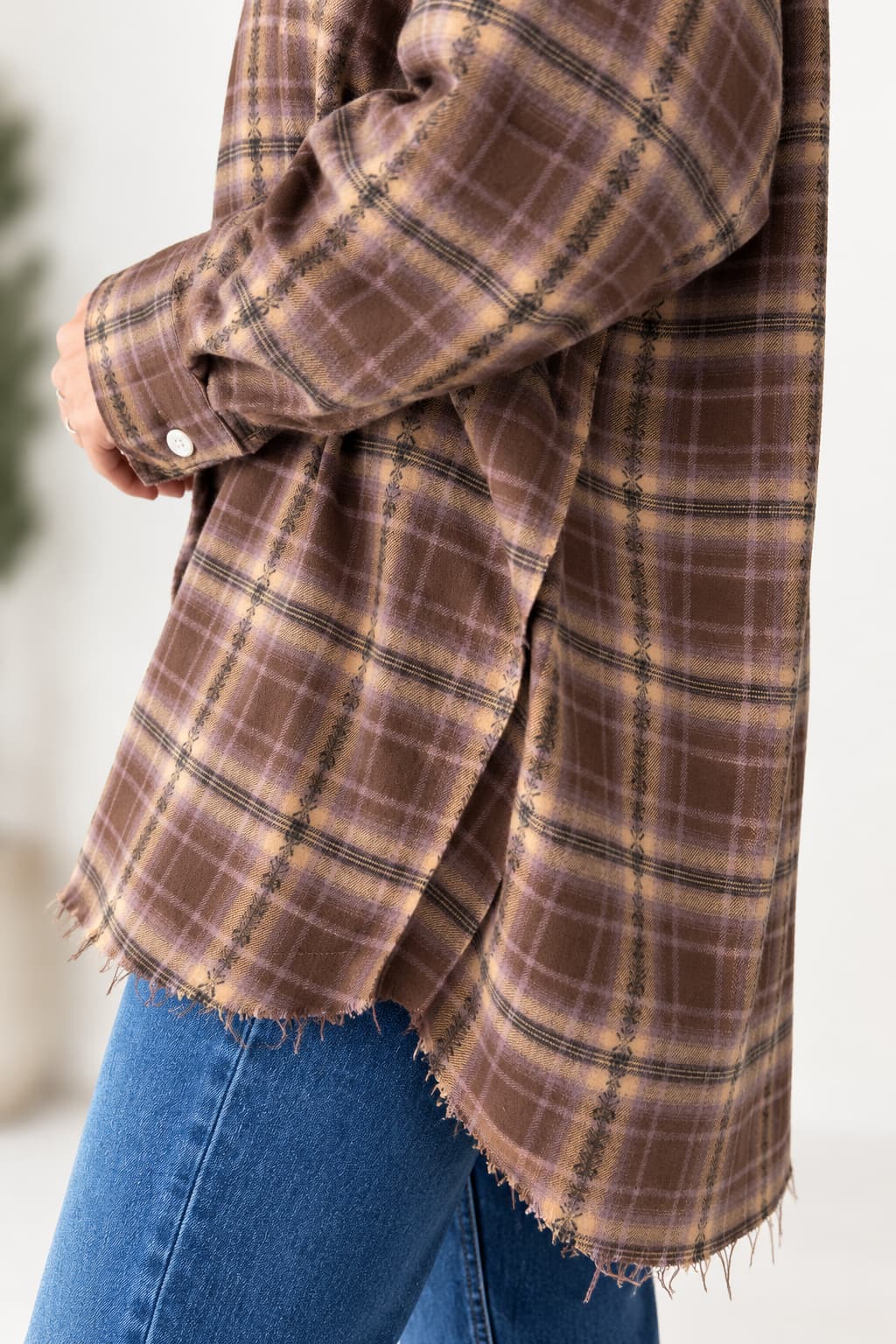 A close-up of a person wearing a brown and beige plaid flannel shirt with a frayed hem, paired with blue jeans.