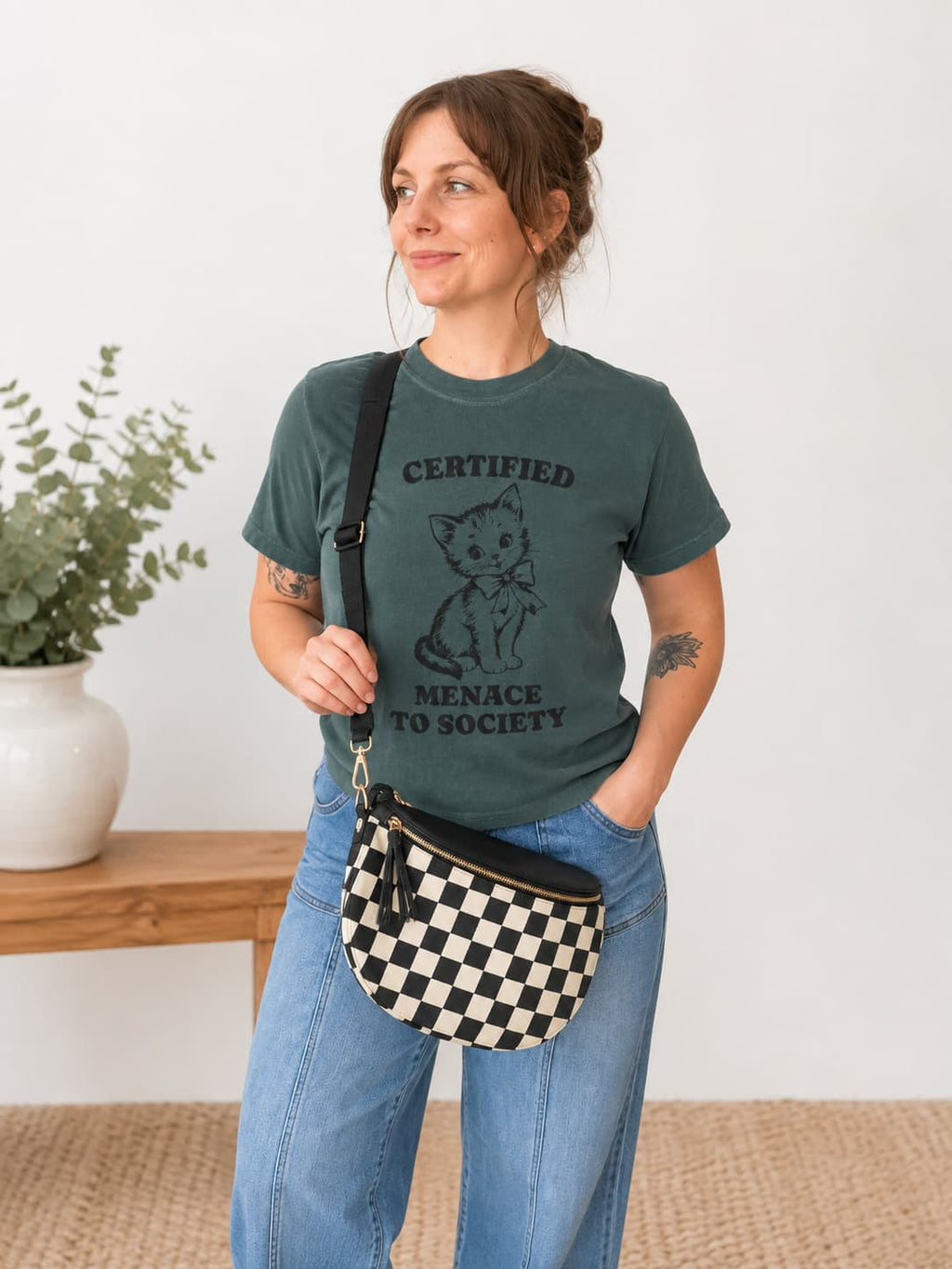 A woman wearing a dark green graphic t-shirt with a black kitten print and the text 'CERTIFIED MENACE TO SOCIETY', paired with blue jeans and a black-and-white checkered crossbody bag, standing in a bright room.