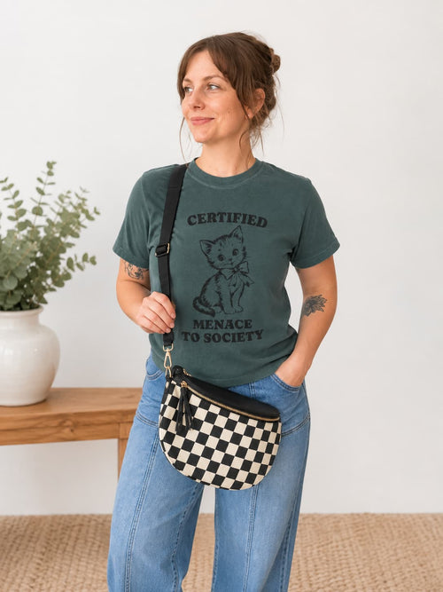 A woman wearing a dark green graphic t-shirt with a black kitten print and the text 'CERTIFIED MENACE TO SOCIETY', paired with blue jeans and a black-and-white checkered crossbody bag, standing in a bright room.