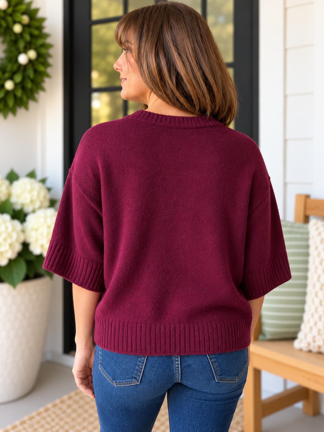 A woman viewed from behind wearing a short-sleeve crewneck knit sweater in deep burgundy, paired with blue jeans, standing on a porch.