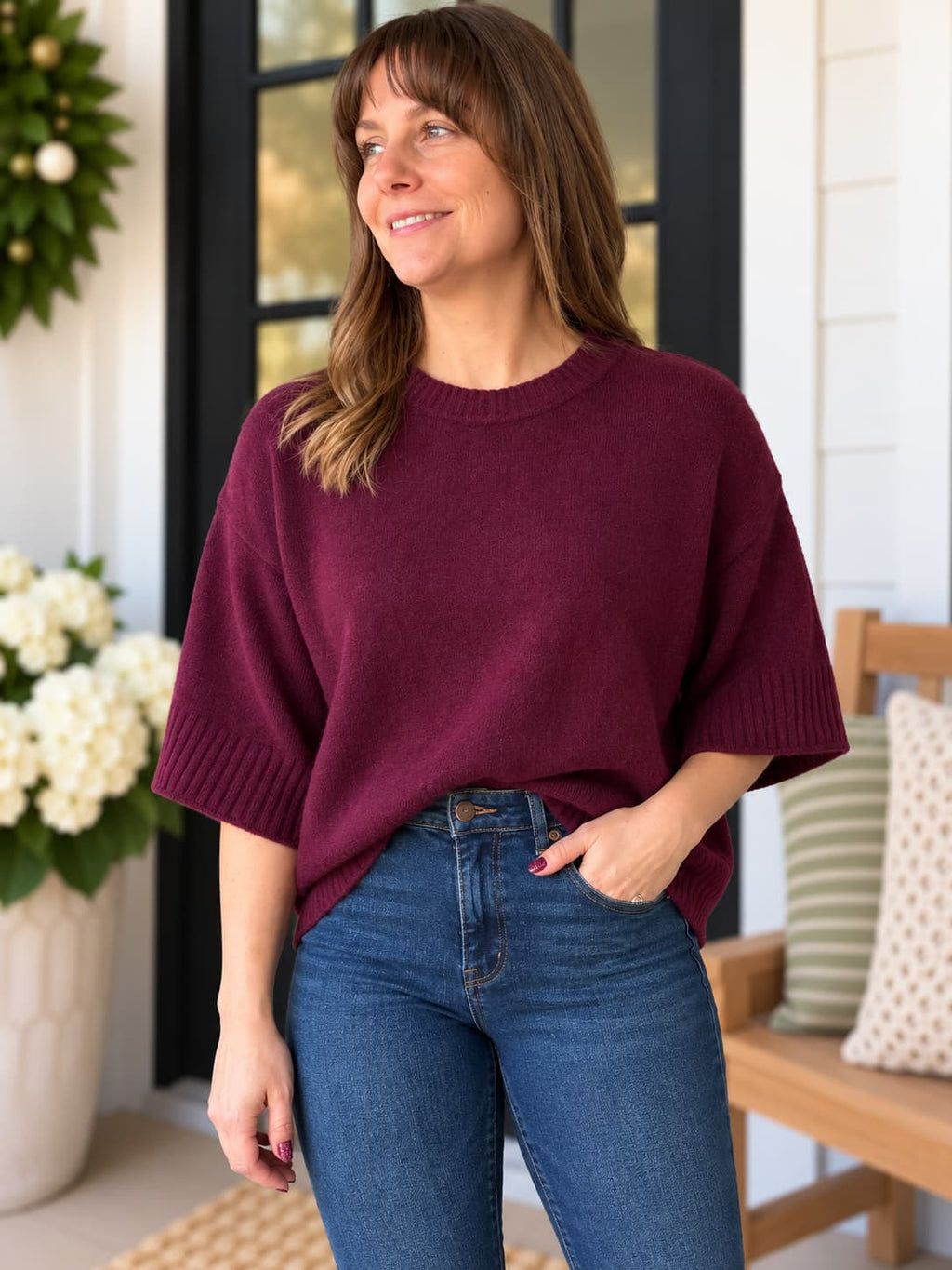 A woman wearing a loose-fitting, short-sleeve crewneck sweater in deep burgundy and blue jeans, standing on a porch with a wooden bench and white flowers in the background.
