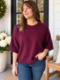 A woman wearing a loose-fitting, short-sleeve crewneck sweater in deep burgundy, paired with blue jeans, standing on a porch.