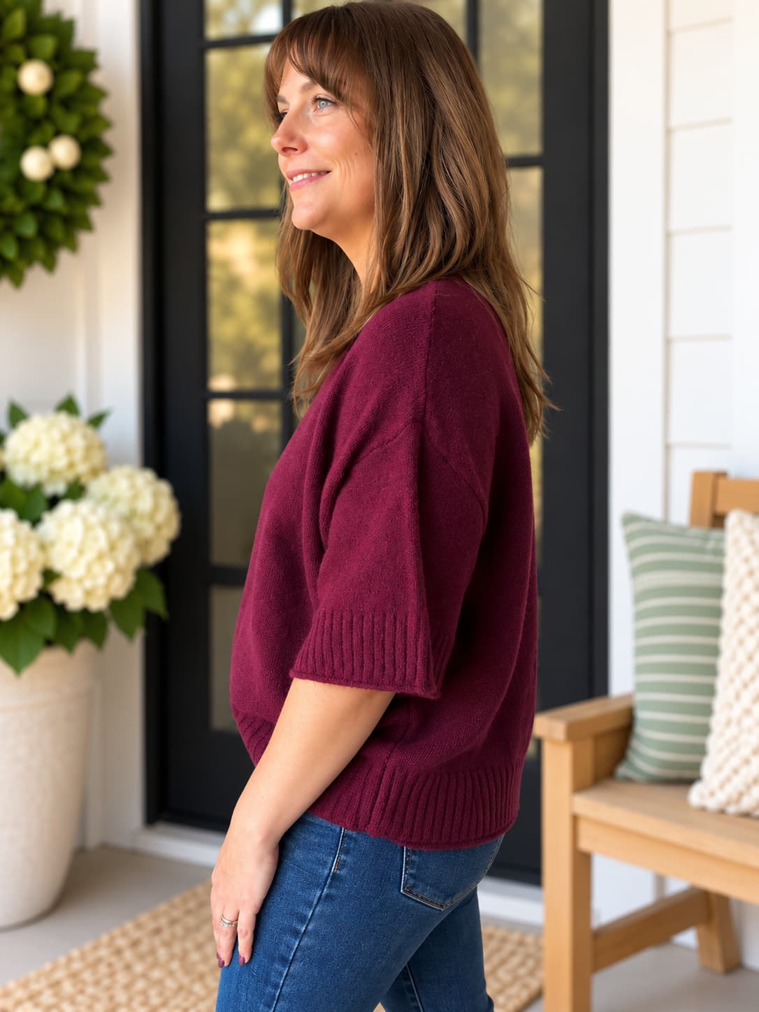 A woman wearing a short-sleeve burgundy knit sweater and blue jeans, standing on a porch in front of a black-framed glass door.