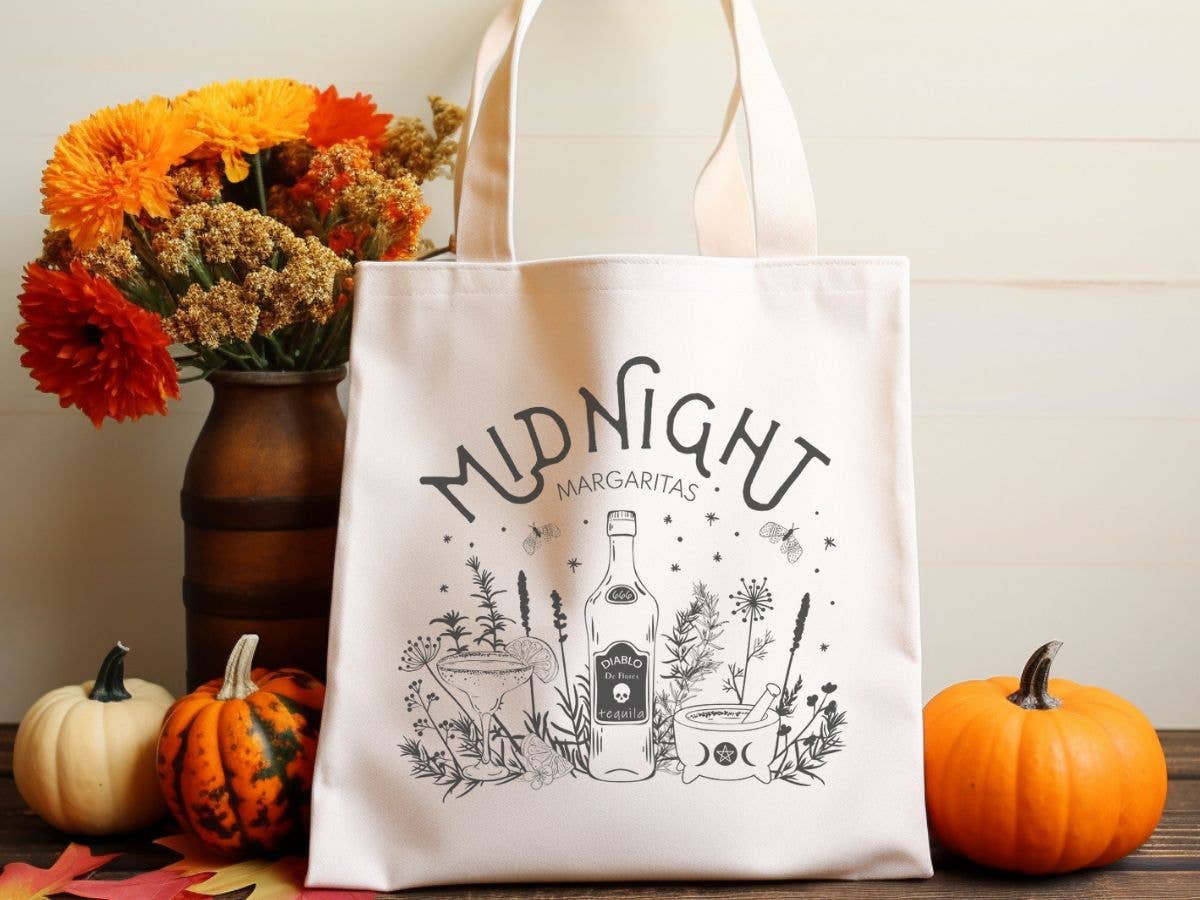 Tote bag with 'Midnight Margaritas' design next to pumpkins and flowers