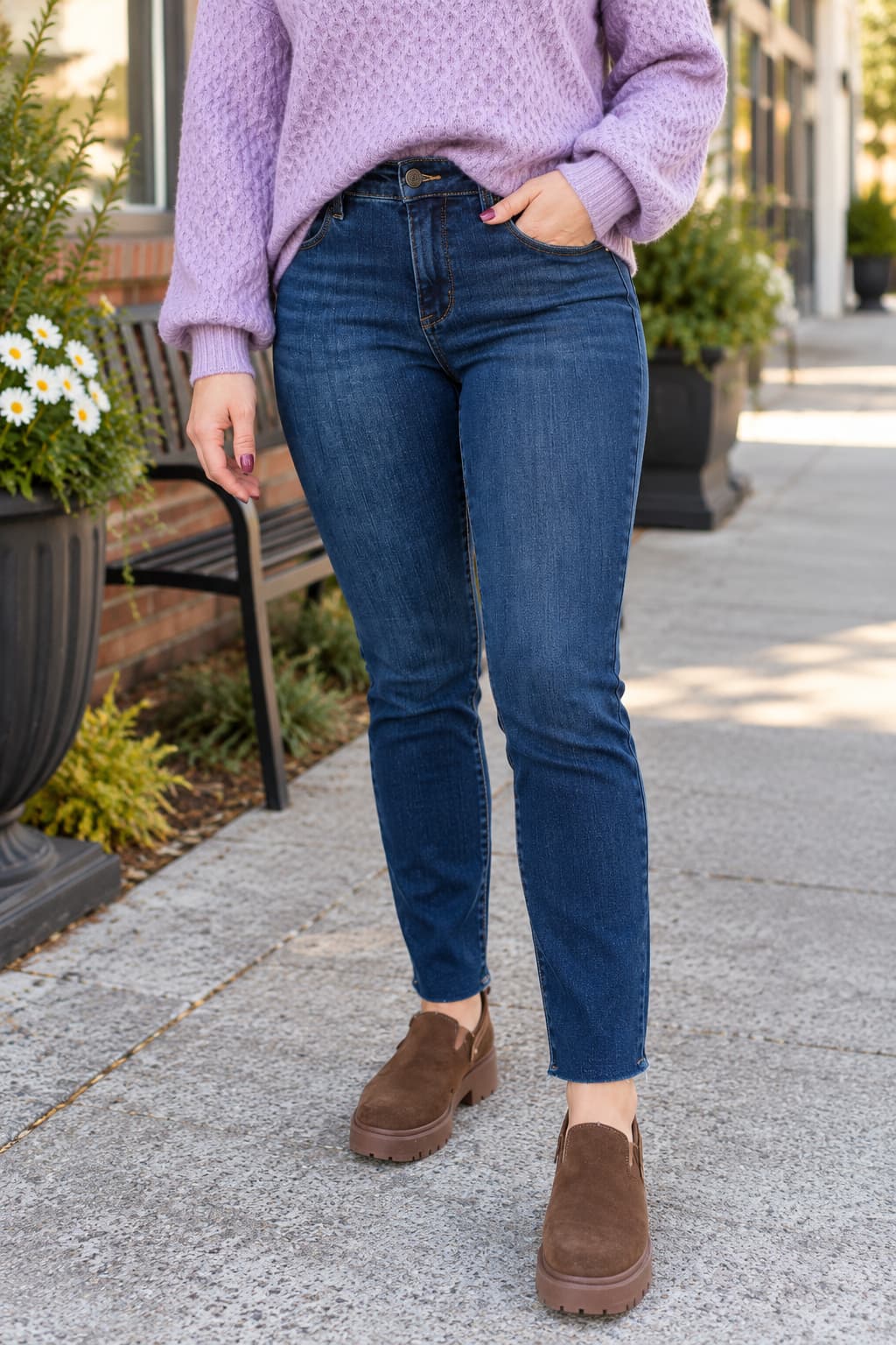 A person wearing dark blue straight leg jeans, a light purple knit sweater, and brown slip-on shoes, standing on a paved outdoor walkway.