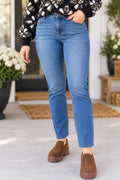 A person wearing light blue slim-fit straight jeans, a black floral long-sleeve top, and brown slip-on loafers, standing on a porch with potted plants in the background.