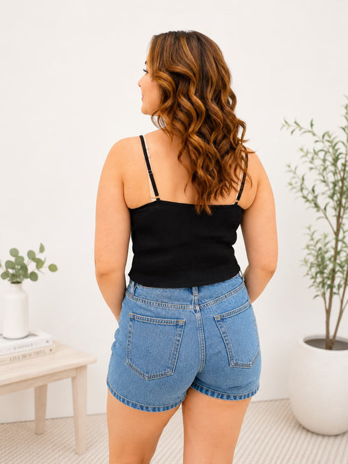 Woman wearing a black tank top and blue denim shorts standing in a room with plants.