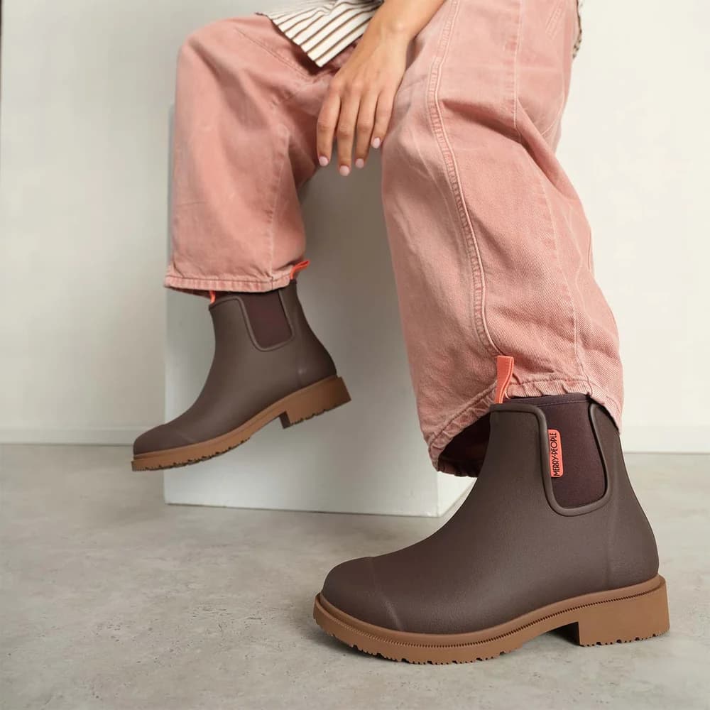 A person seated on a white block wearing dark brown Chelsea-style rain boots with elastic side panels and tan soles, paired with loose pink pants.