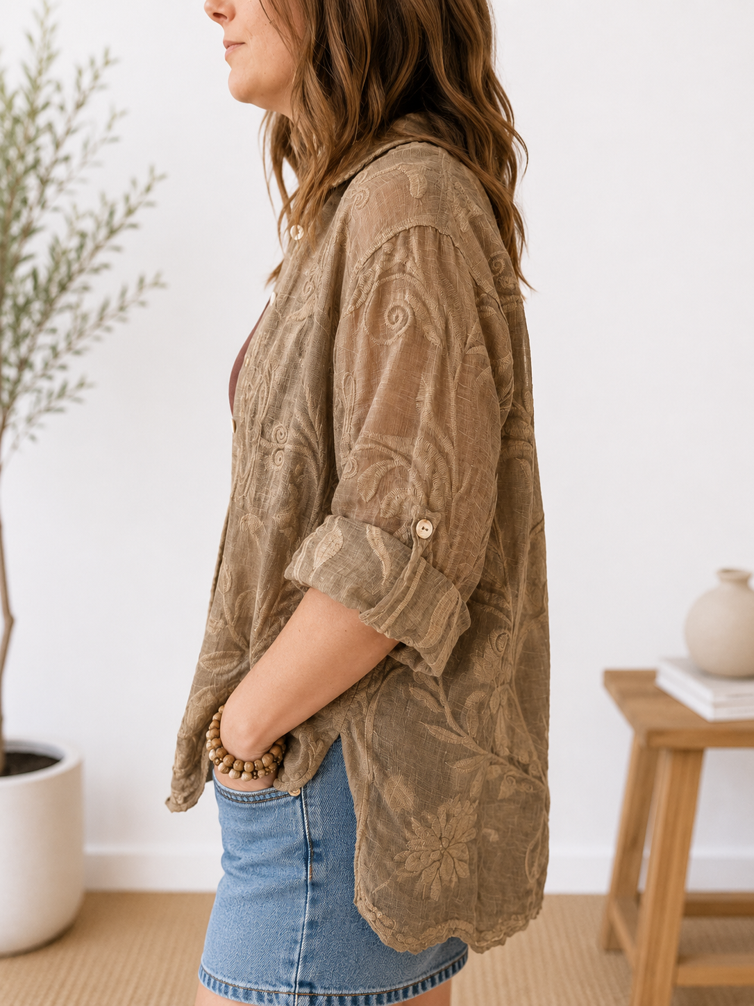 A woman shown in side profile wearing a loose, semi-sheer taupe-brown button-up shirt with a tonal floral pattern, paired with a denim skirt in a bright, modern room.