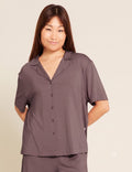 A woman modeling a short-sleeve button-front pajama top in a muted taupe-brown color, paired with matching pants, against a plain light beige studio background.