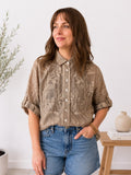 A woman wearing a short-sleeve button-up shirt with a brown and beige damask pattern, paired with blue denim shorts, standing in a brightly lit room.