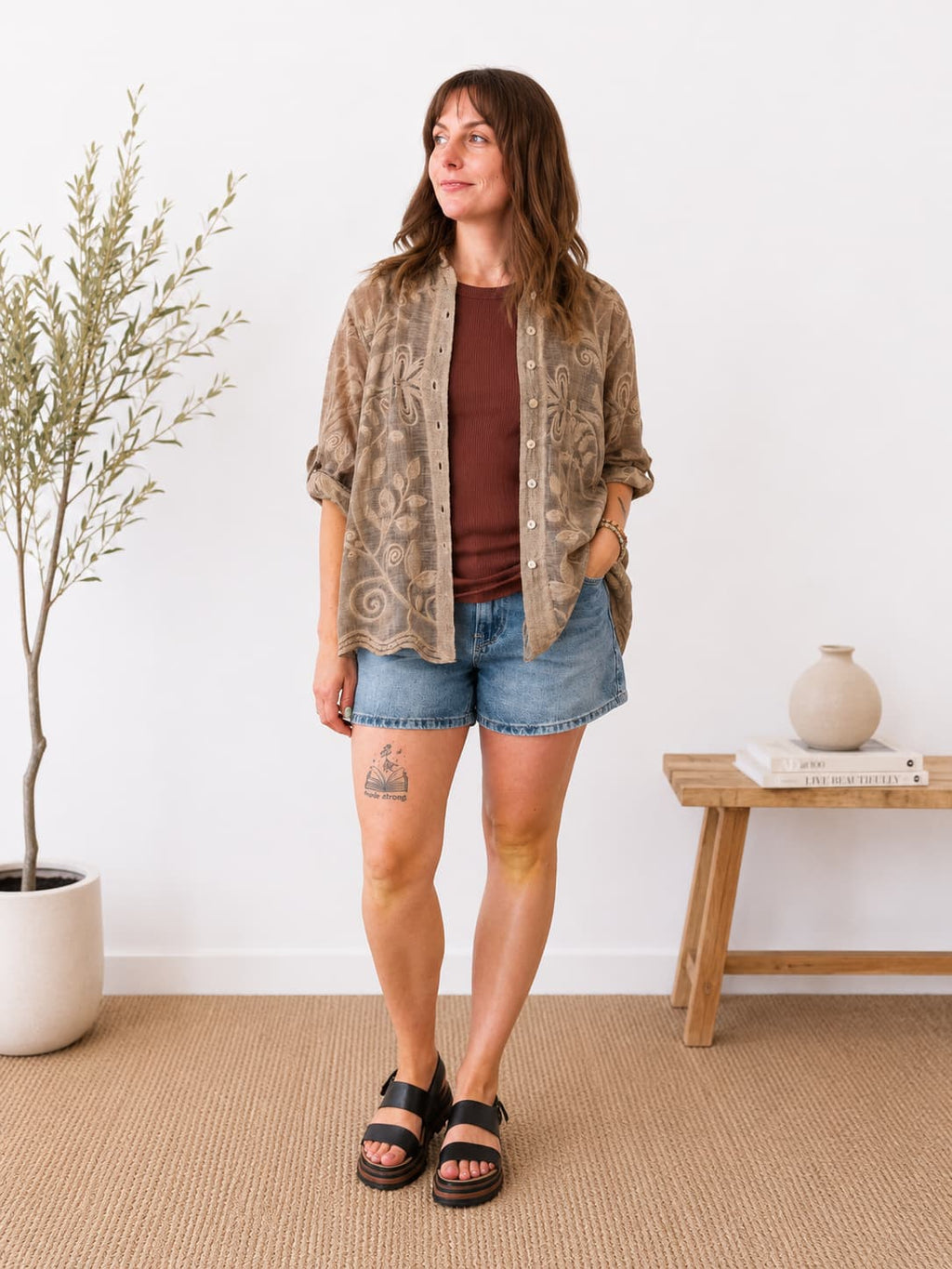 A woman stands in a bright room wearing a brown bohemian patterned button-up shirt over a rust-brown t-shirt, paired with denim shorts and black slide sandals.