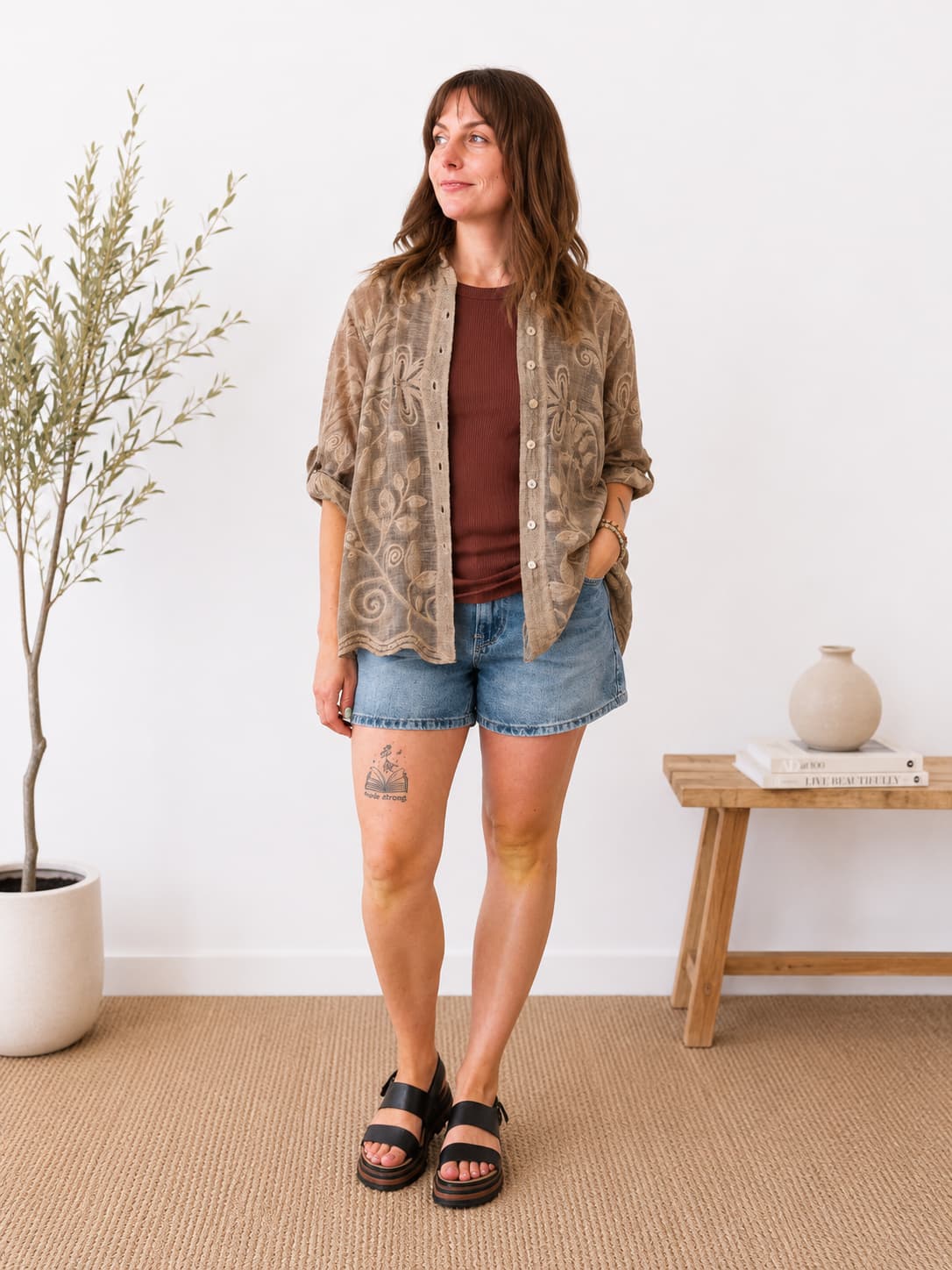 A woman stands in a bright room wearing a brown bohemian patterned button-up shirt over a rust-brown t-shirt, paired with denim shorts and black slide sandals.